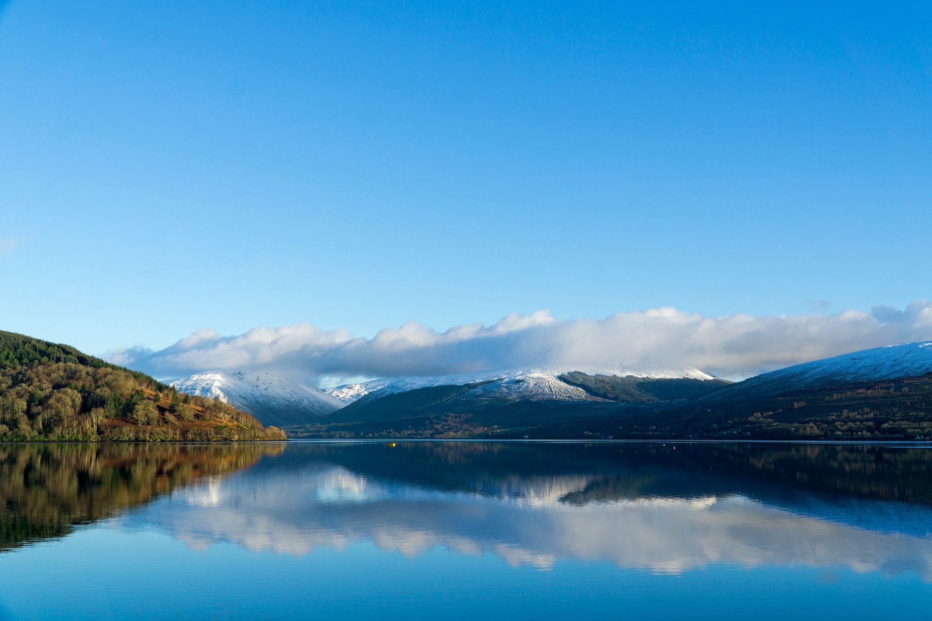 Serene alpine lake with mirror-like reflection of snow-capped mountains and forested slopes under clear blue sky