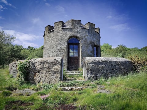 Stone castle tower with crenellated top nestled among boulders and green vegetation under blue sky